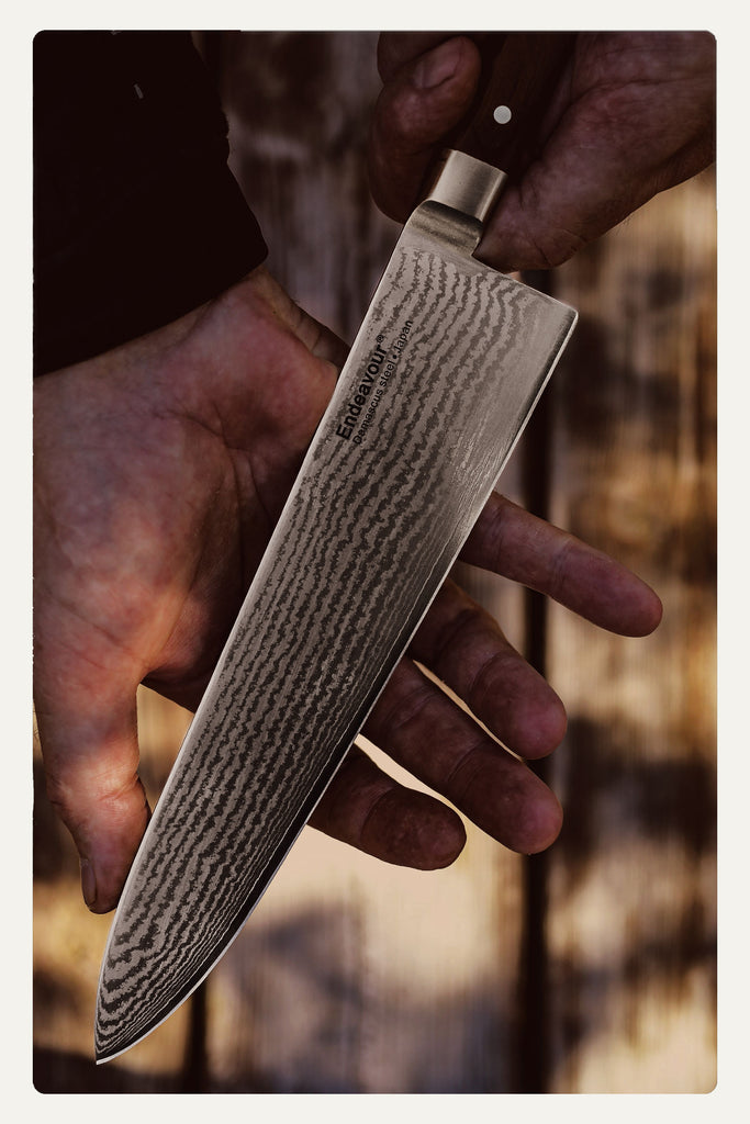  A hand holds a large chef's knife with a patterned blade against a wooden background.